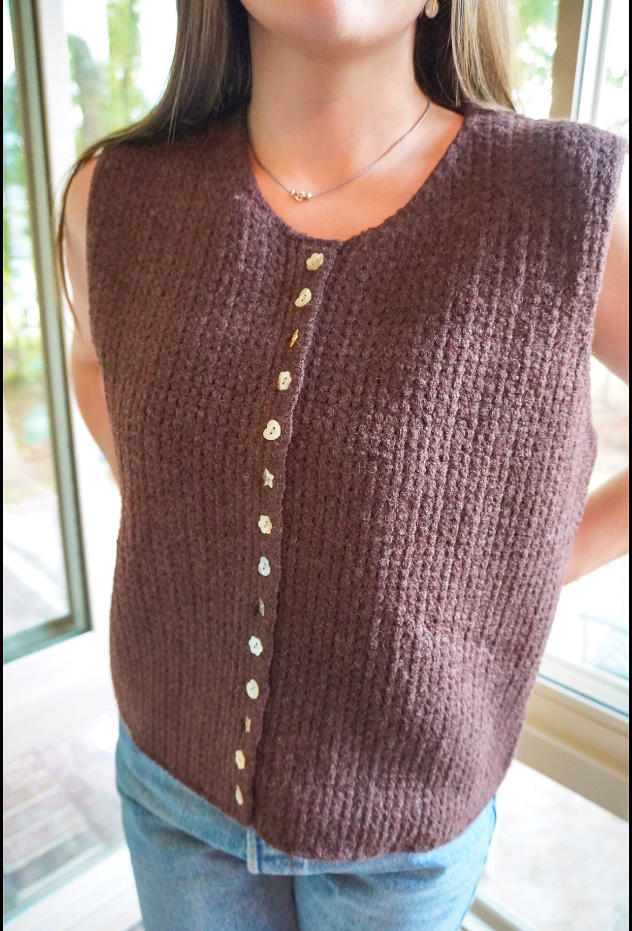 A woman wearing a sleeveless, ribbed knit sweater vest in a dark brownish-purple color, featuring a vertical row of light-colored buttons down the front. She is also wearing blue jeans and a delicate necklace, standing in front of a window.
