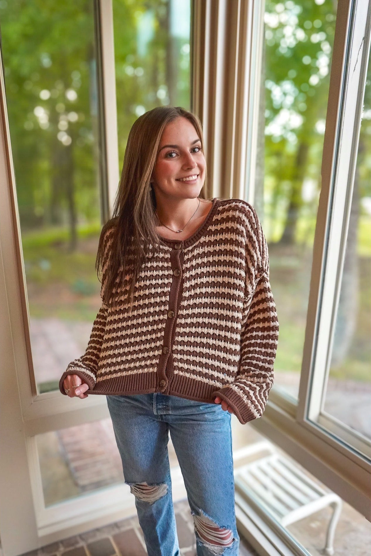 A woman wearing a brown and beige patterned knit cardigan sweater and ripped blue jeans, standing in front of large windows with a green outdoor background.
