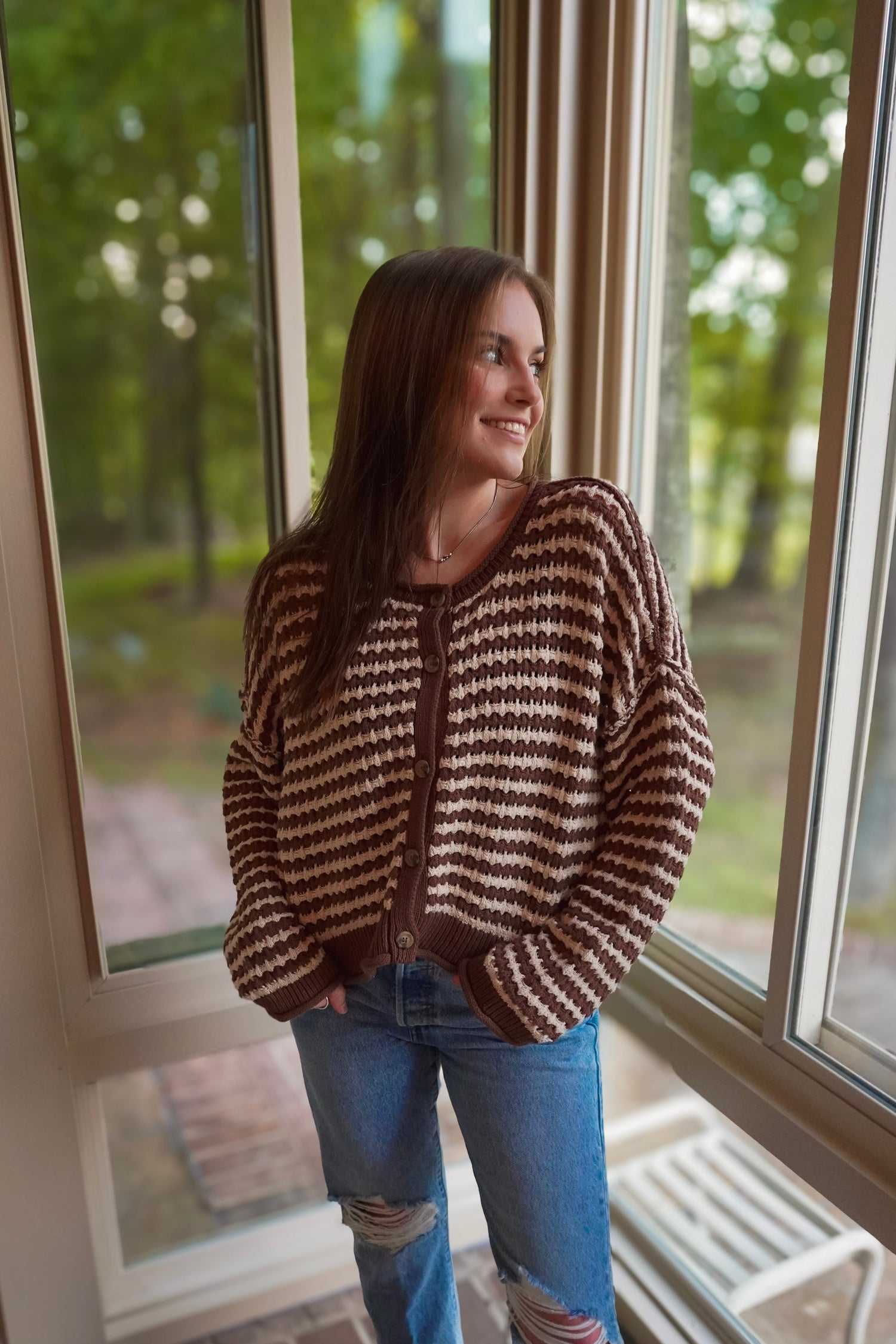 A woman wearing a brown and beige striped knit cardigan with a brown zipper, paired with blue jeans, standing in front of large windows with a blurred outdoor forest background.