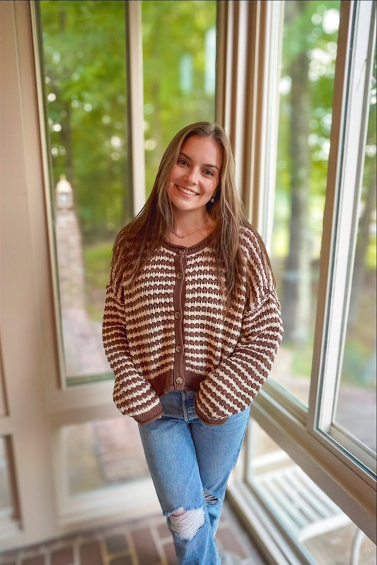 A woman wearing a brown and cream patterned knit cardigan with blue jeans, standing in front of large windows with a blurred outdoor background.