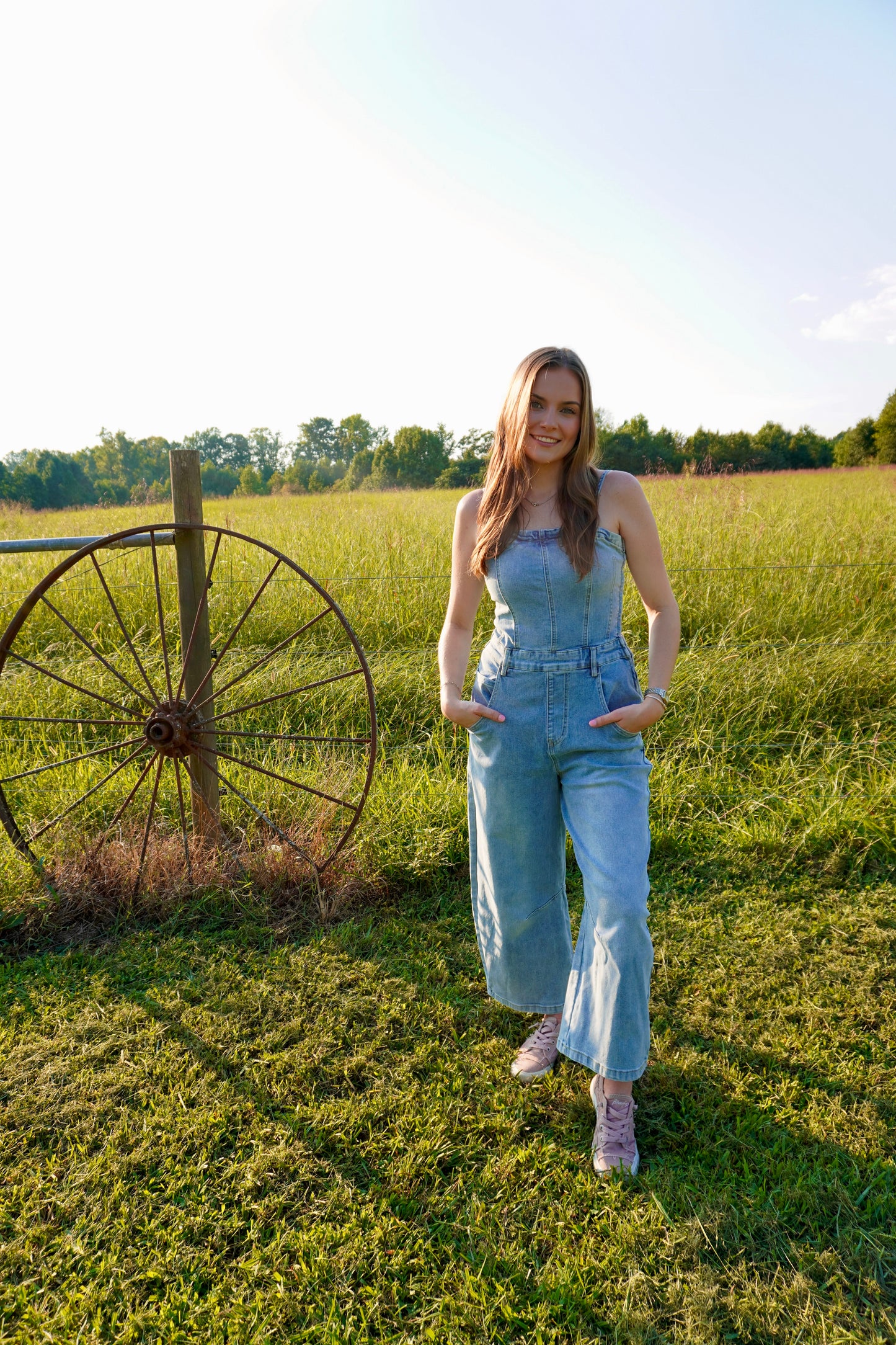 A woman wearing a light blue denim jumpsuit stands in a grassy field next to a large vintage wagon wheel. The background features a bright sky and a line of trees in the distance.