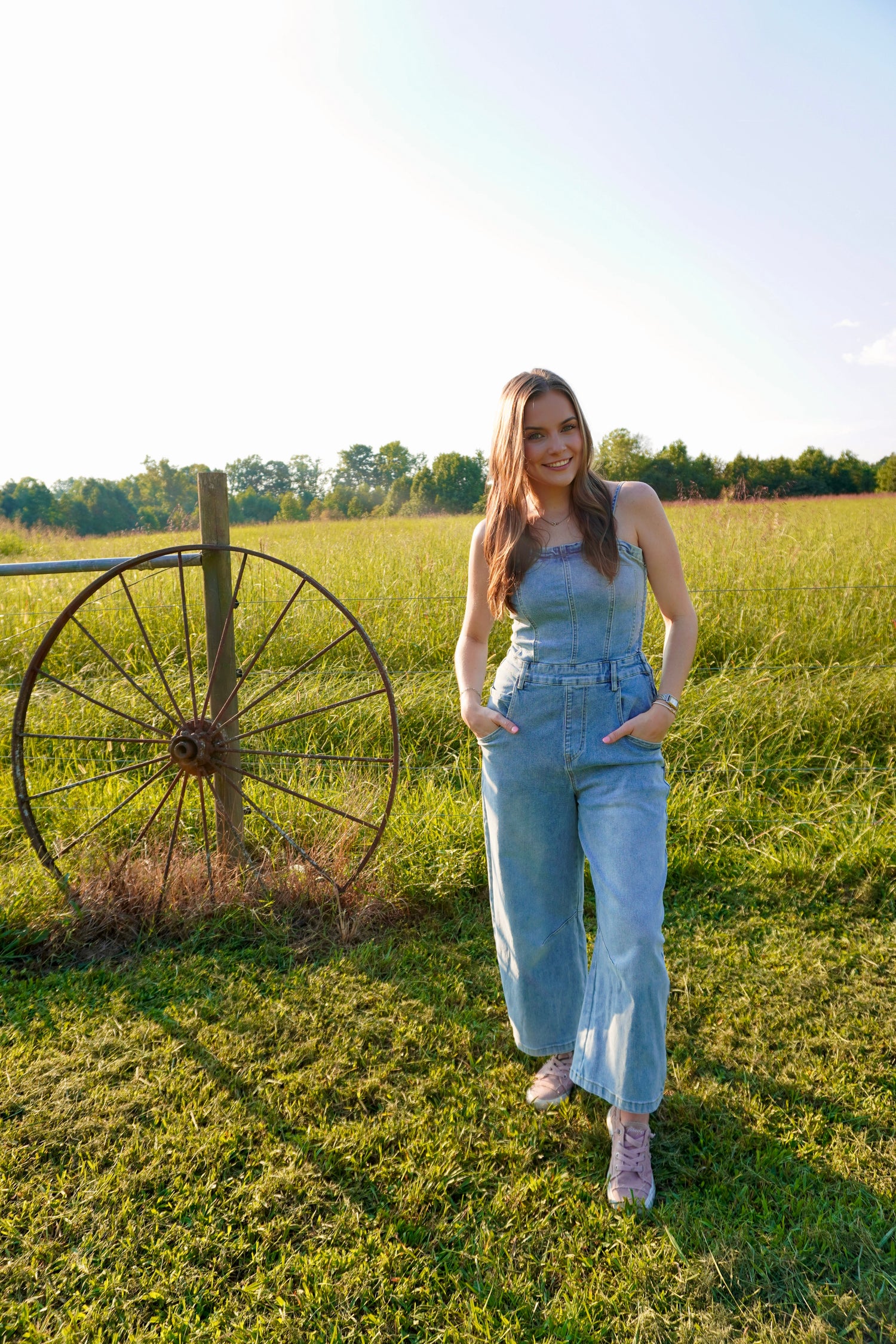 A woman stands in a grassy field wearing a light blue denim overall jumpsuit, with a rustic wooden fence and large metal wagon wheel in the background.