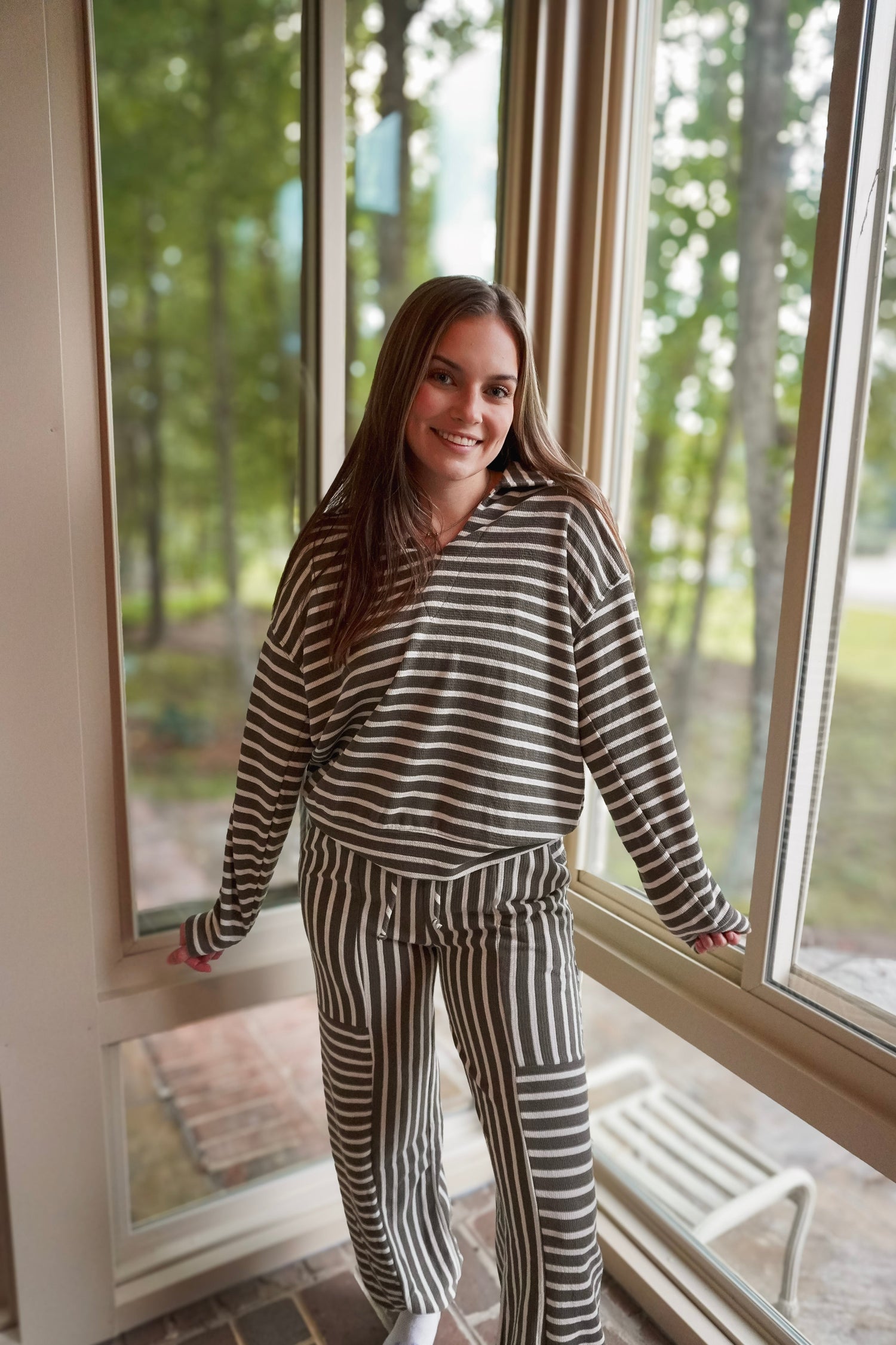 A woman wearing a matching two-piece loungewear set with horizontal black and white stripes, standing in a sunroom with large windows looking out onto a green wooded background.