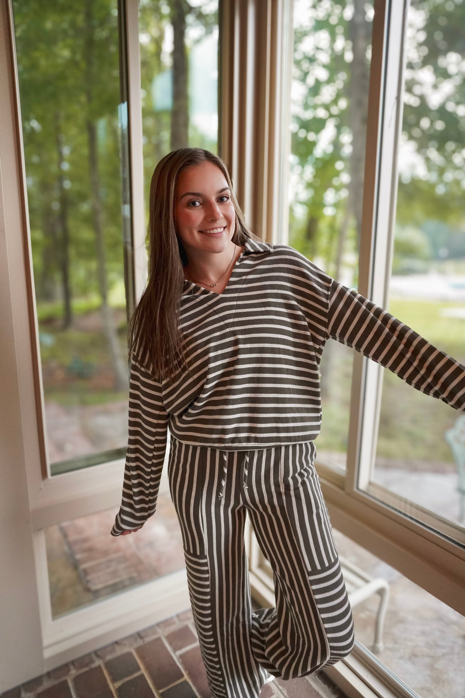 A woman wearing a matching two-piece loungewear set with dark brown and beige horizontal and vertical stripes, standing in a sunroom with large windows looking out onto a green yard.