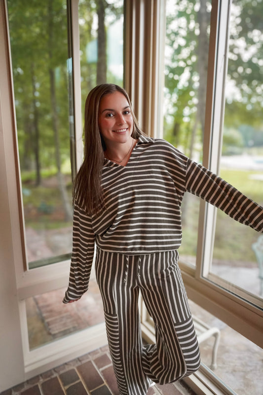 A woman wearing a matching two-piece loungewear set with dark brown and beige horizontal and vertical stripes, standing in a sunroom with large windows looking out onto a green yard.