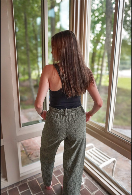 A woman viewed from behind wearing loose, high-waisted olive green patterned pants and a black tank top, standing on a brick porch in front of large windows looking out onto a wooded background.