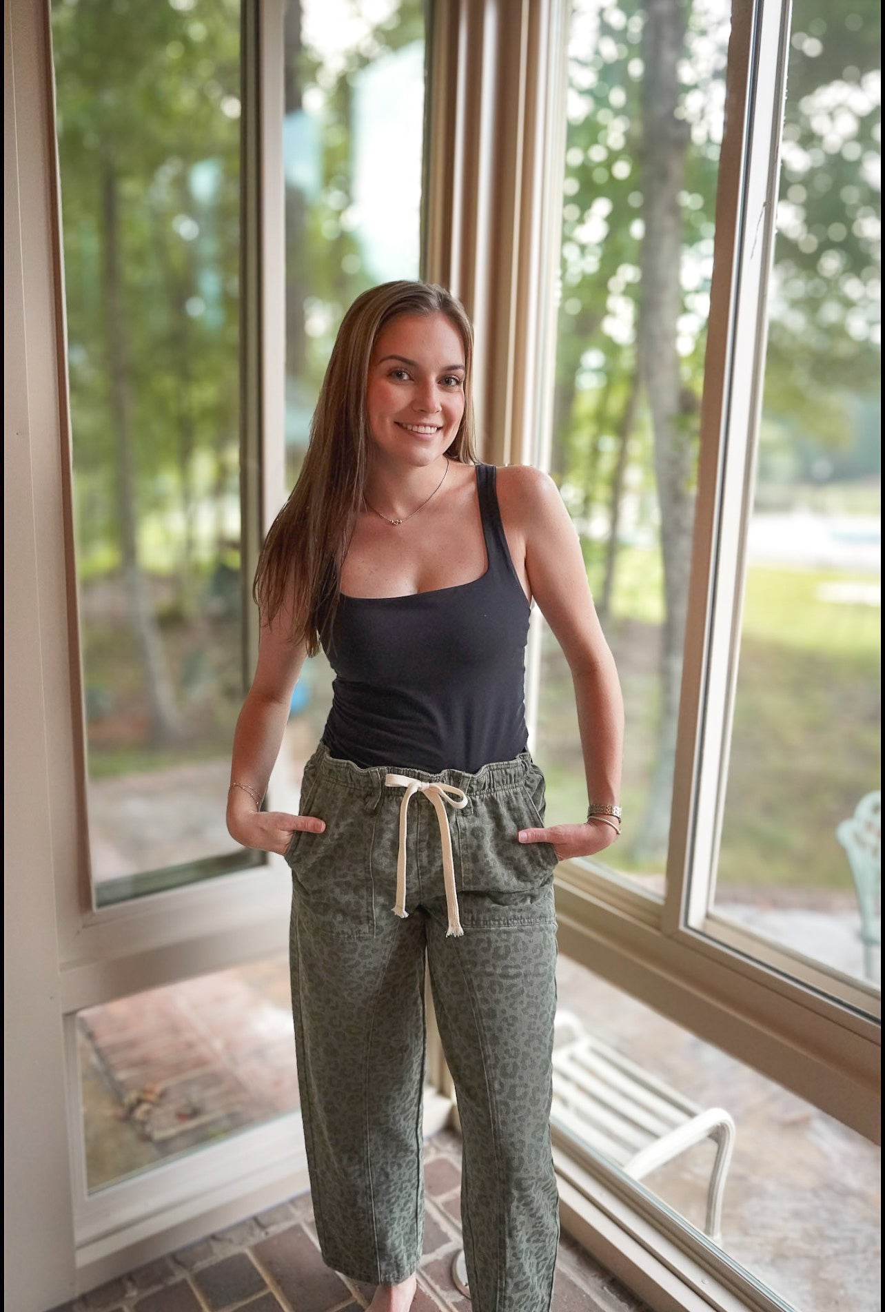 A woman stands in a sunroom wearing loose, relaxed-fit pants in a textured sage green fabric with a cream drawstring, paired with a black tank top.