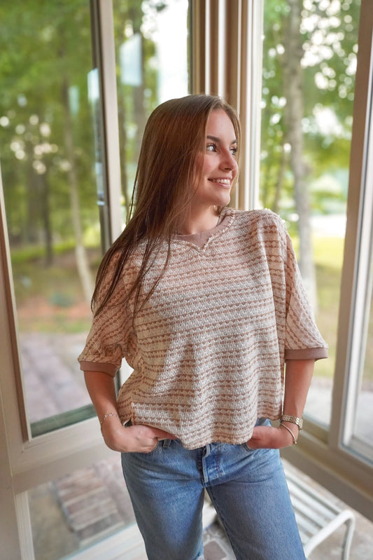 A woman with long light brown hair stands in front of large windows wearing a beige and brown patterned short-sleeve top and blue jeans.