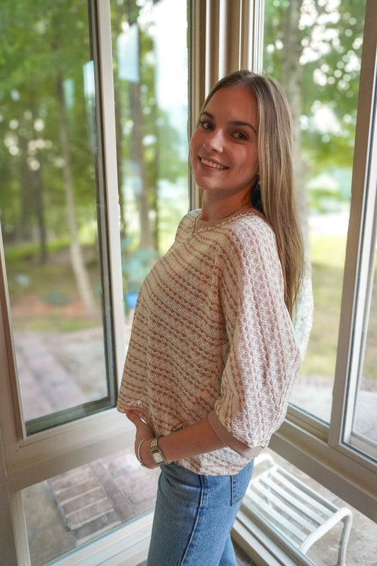 A woman with long blonde hair stands by large windows wearing a cream and brown textured knit top and blue jeans, with a blurred outdoor background of trees and a bench.
