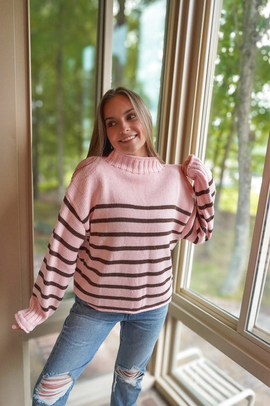 A woman wearing a pink knit sweater with dark brown horizontal stripes and light blue ripped jeans, standing in front of a large window with a blurred outdoor background.