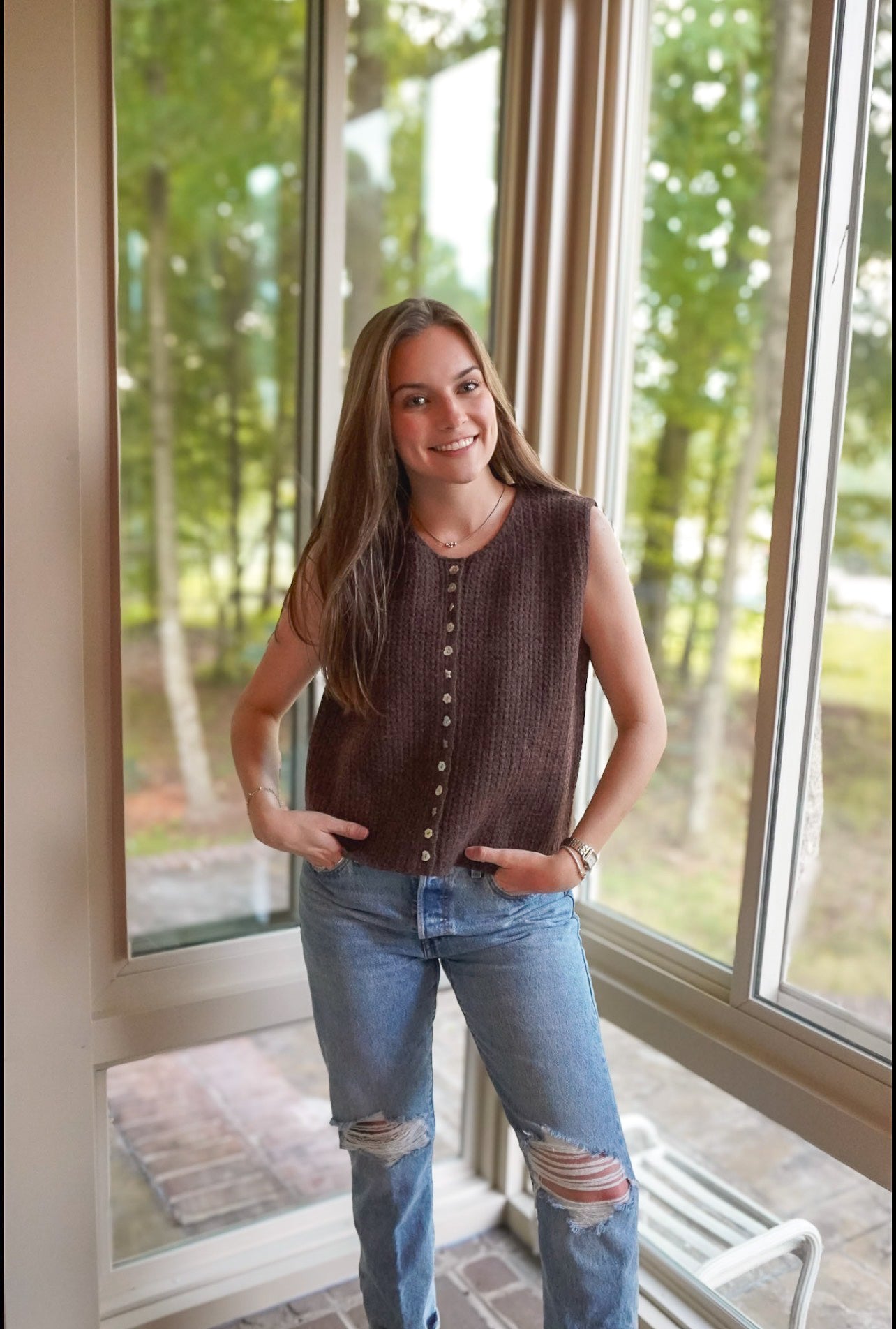 A woman stands indoors by large windows wearing a sleeveless brown knit top with a button front and light blue ripped jeans.