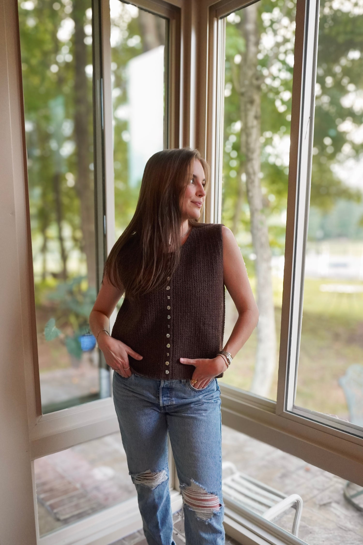 A woman wearing a dark brown sleeveless knit vest with front buttons and light blue ripped jeans, standing in a sunroom by large windows looking out onto a wooded backyard.