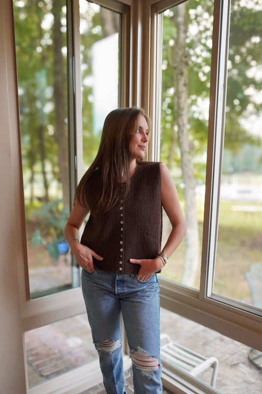 A woman wearing a dark brown sleeveless knit vest with front buttons and light blue ripped jeans, standing in a sunroom by large windows looking out onto a wooded backyard.
